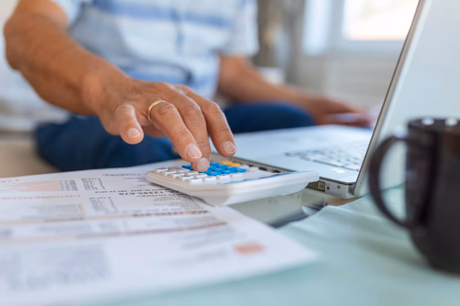 Hand using calculator next to laptop and financial documents on a glass table.