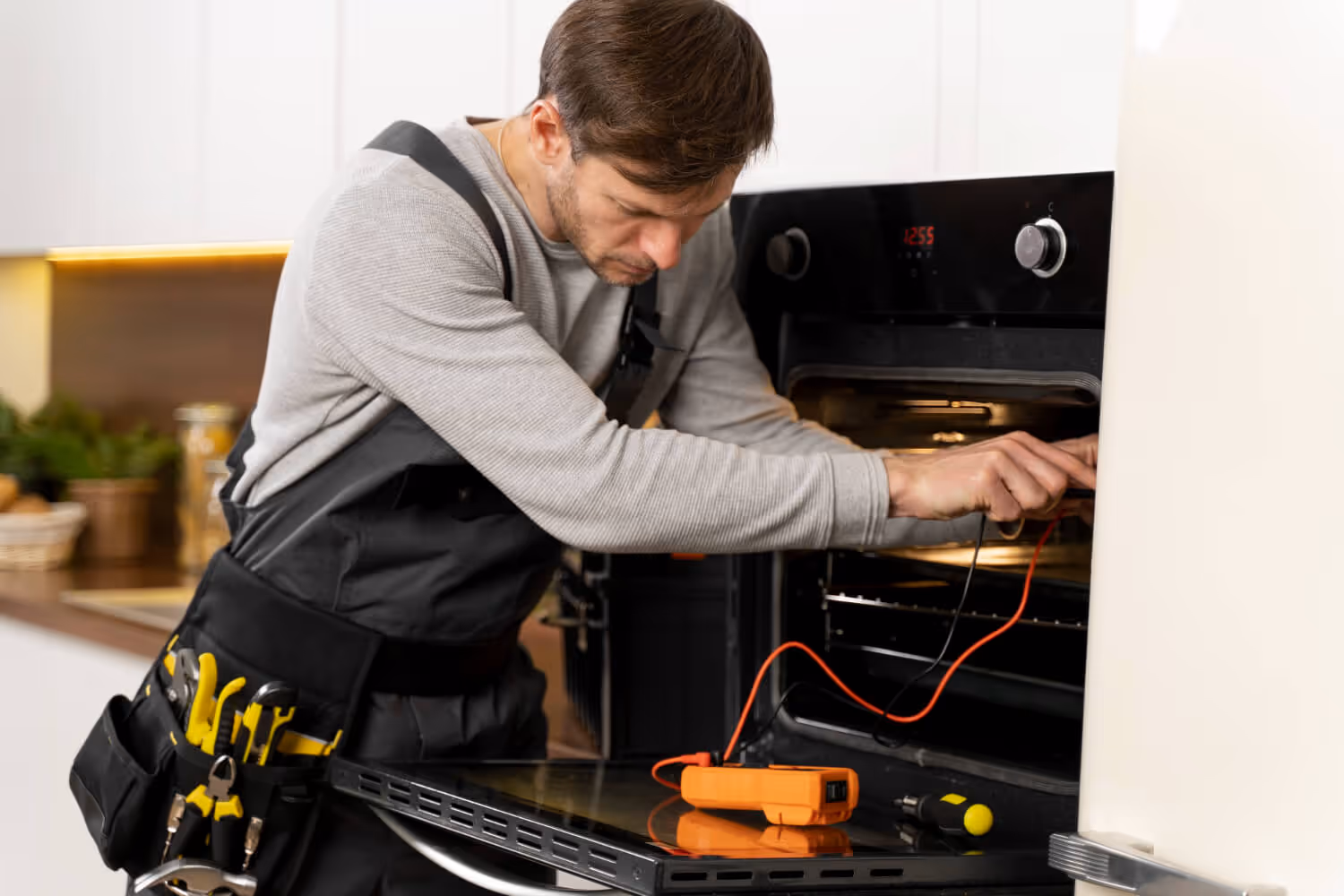 Technician in grey shirt and black overalls testing an oven with an orange multimeter.
