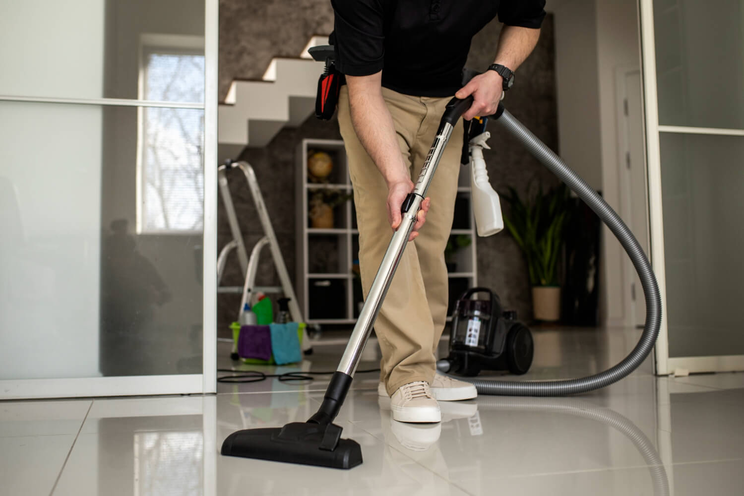 Person vacuuming a shiny tiled floor indoors with cleaning supplies and a ladder in the background.