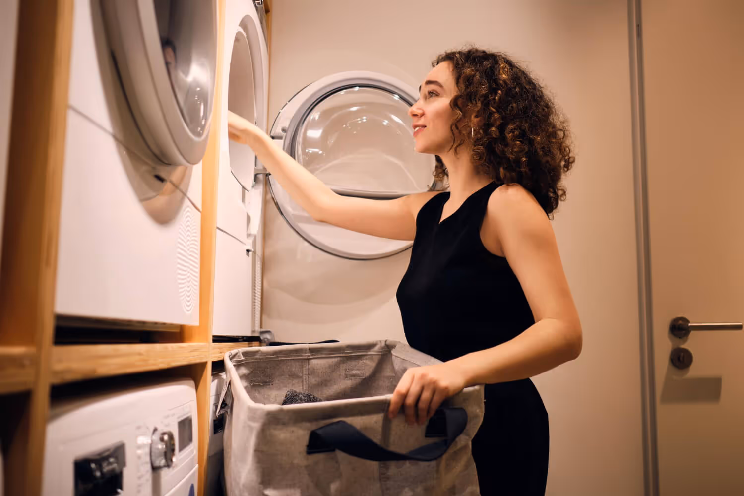 Woman with curly hair putting clothes into a front-loading washing machine in a laundry room.
