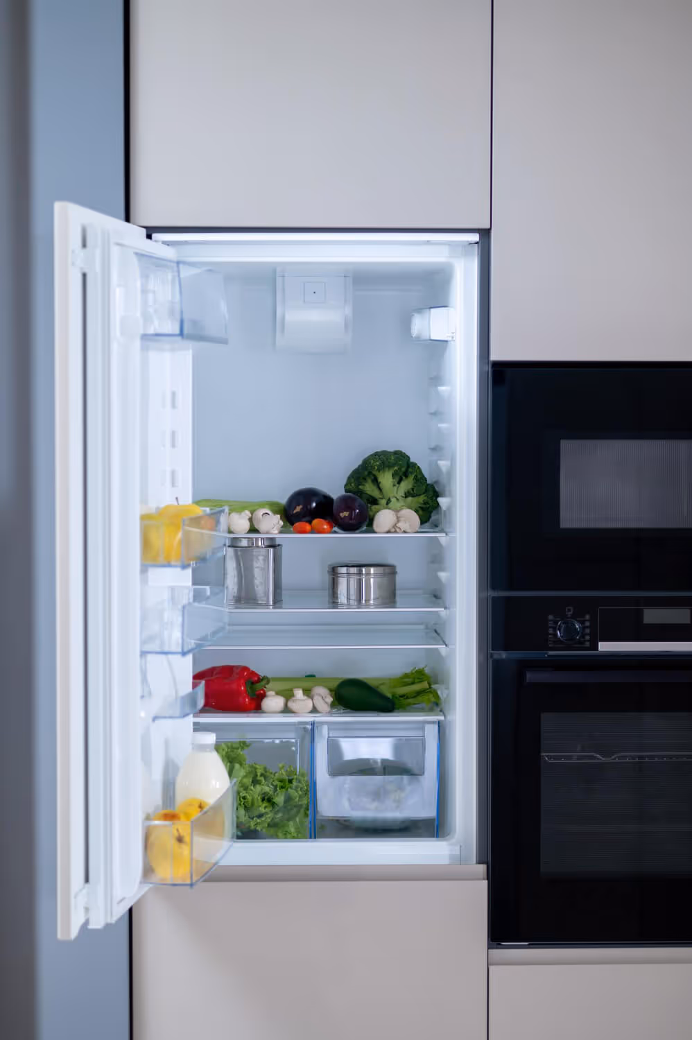 Open refrigerator filled with various vegetables including broccoli, eggplants, mushrooms, cherry tomatoes, lettuce, celery, cucumber, and a bottle of milk on the door shelf.
