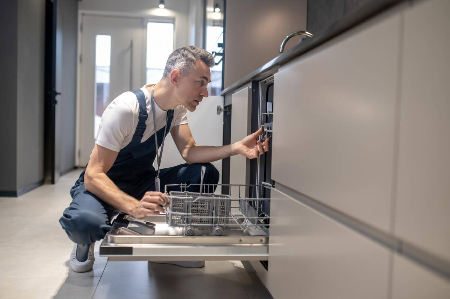Repair technician inspecting the inside mechanism of an open dishwasher in a modern kitchen.