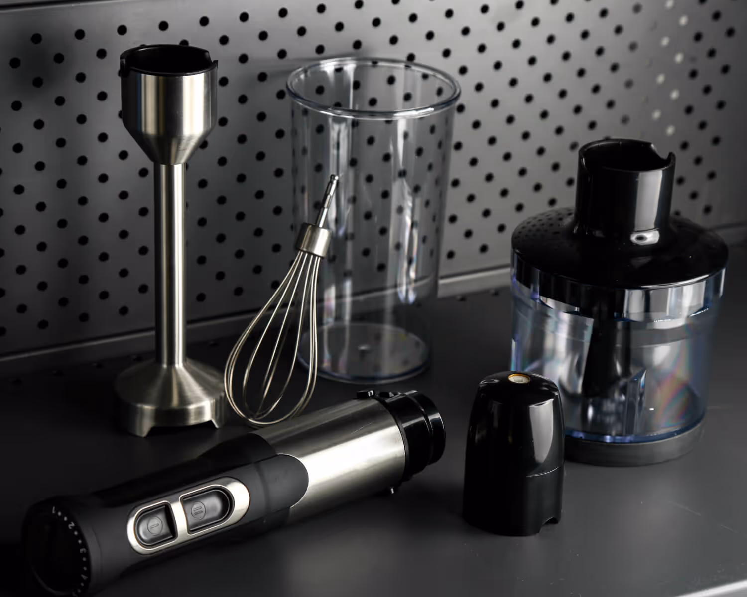 Disassembled black and stainless steel hand blender with whisk, blending shaft, and plastic container on a gray surface against a perforated metal wall.