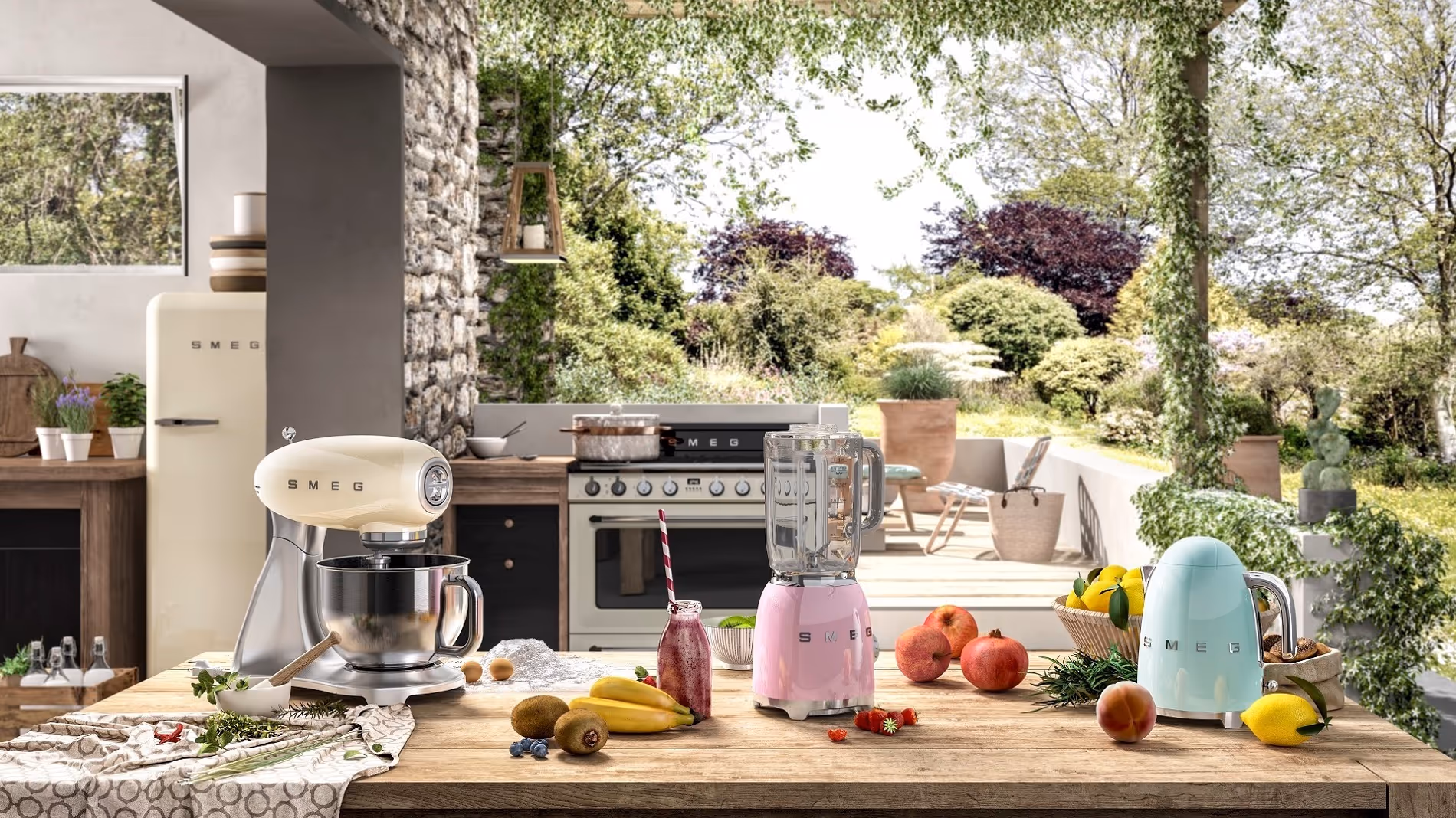 Wooden kitchen table with pastel SMEG mixer, pink blender, turquoise kettle, fresh fruits, and herbs in a bright kitchen opening to lush garden.