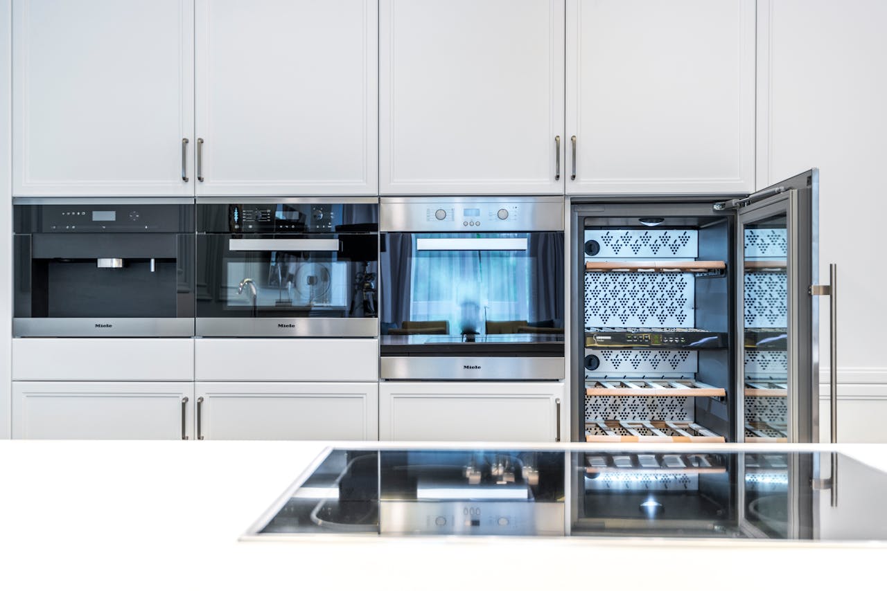 Modern built-in kitchen appliances with a coffee machine, oven, and open wine cooler with wooden racks in white cabinetry.